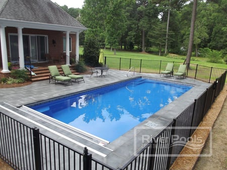 Blue rectangular fiberglass pool with grey patio and green trees around it.