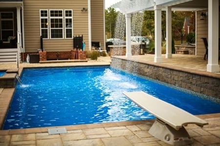 T40 pool in Maya Blue with diving board, waterfall off pergola, elevated tanning ledge, and contrasting tan/brown patio and coping