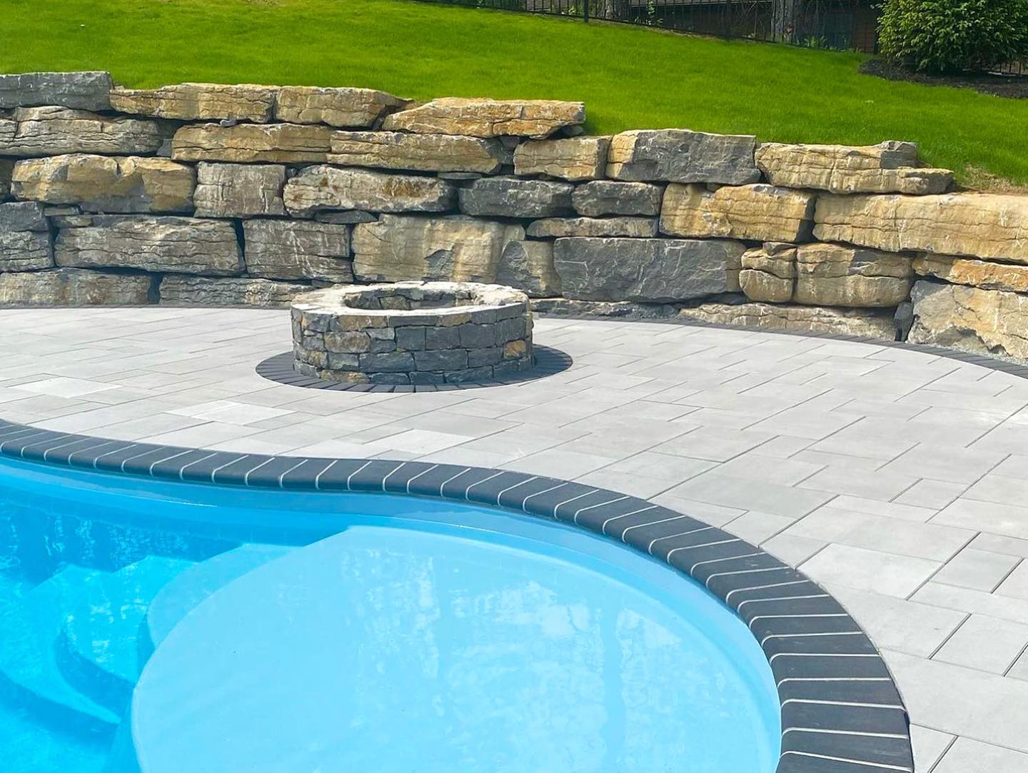 Close-up of curved fiberglass pool with stone fire pit and retaining wall in a backyard installation in upstate NY