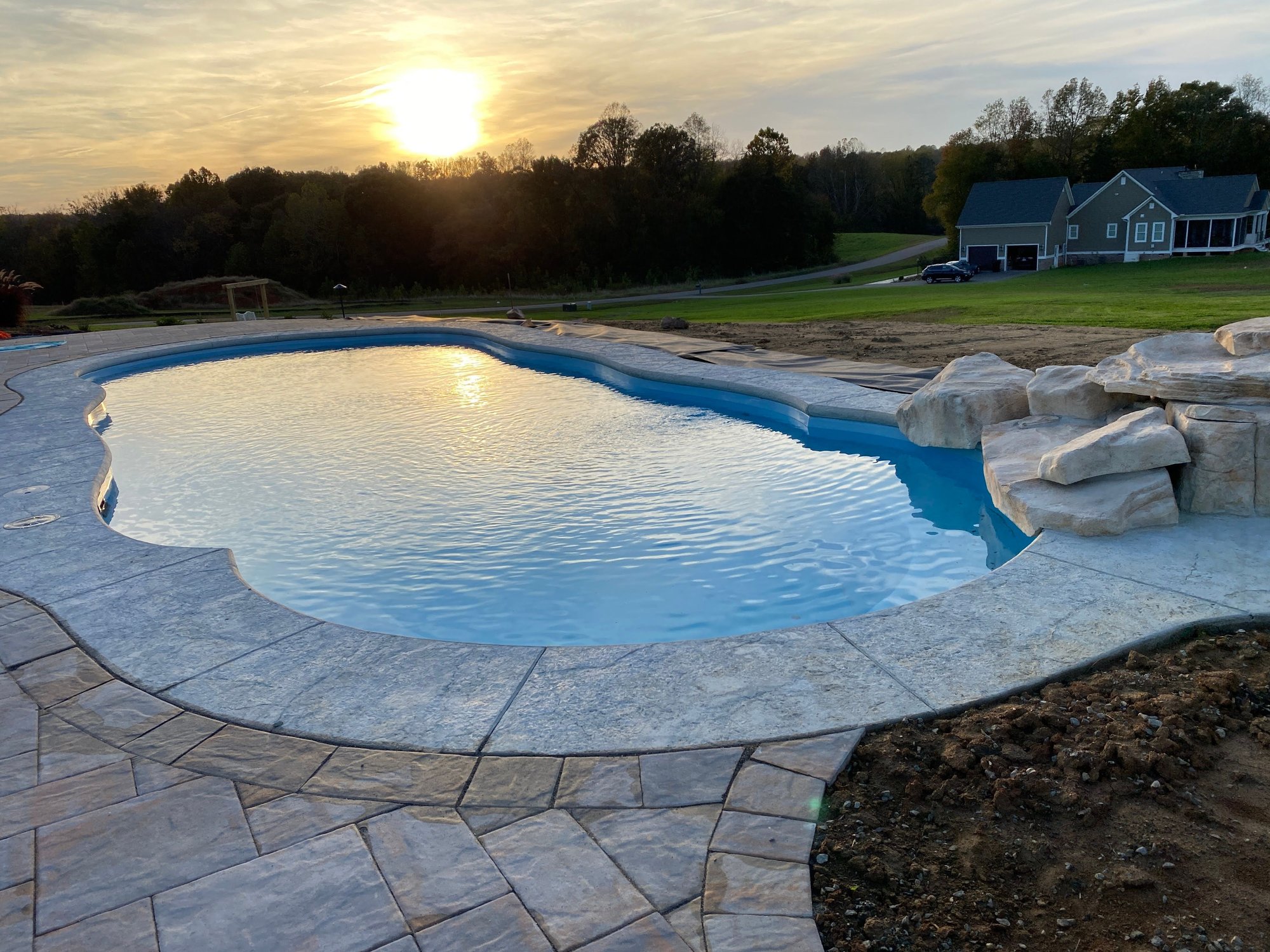 Fiberglass inground pool installation in Rogers, AR, with stamped concrete patio, natural rock accents, and sunset view.