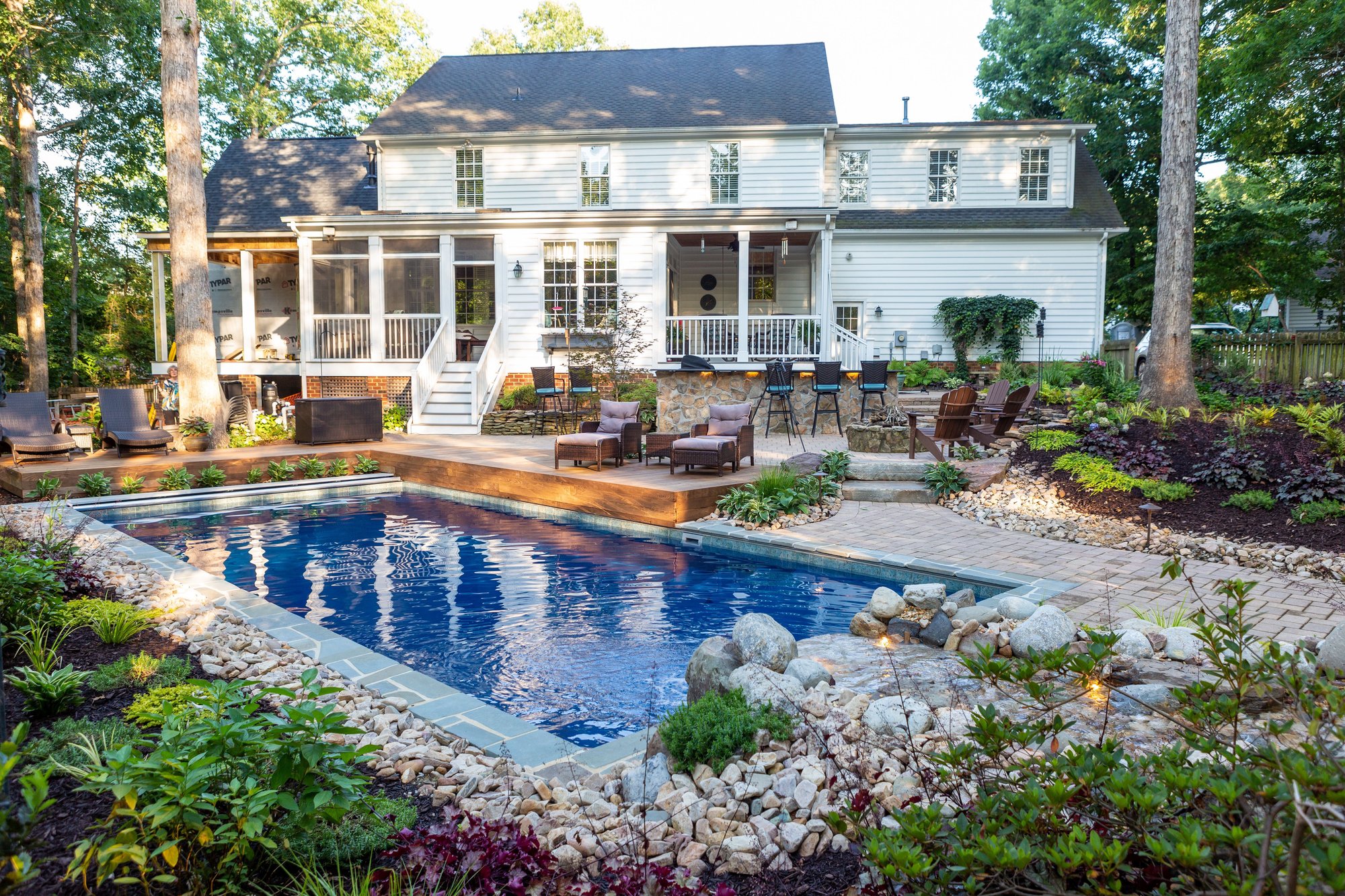 Fiberglass inground pool installation in Rogers, MO, with natural stone accents, paver decking, wood deck, and lush landscaping.