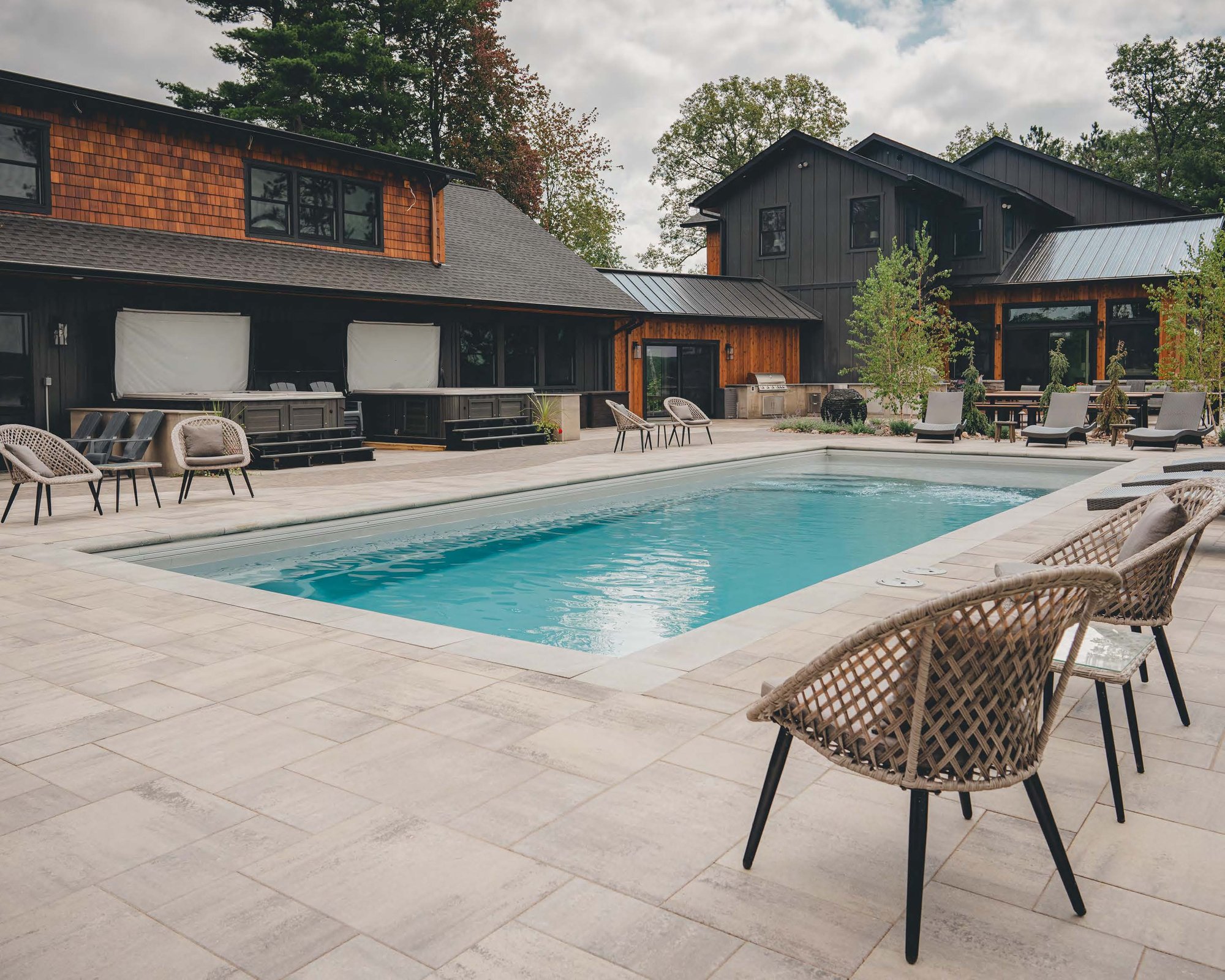 Modern backyard with a rectangular inground fiberglass pool installed by River Pools of Red Wing near Eau Claire, WI, surrounded by large-format pavers, contemporary patio furniture, and a dark-toned farmhouse-style home.
