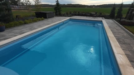 Wide-angle view of a modern fiberglass pool with an automatic pool cover system, surrounded by paver decking and scenic countryside—installed by River Pools Red Wing.