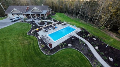 Aerial view of a fiberglass pool installed by River Pools Red Wing, featuring a custom patio, fire pit, and landscaped backyard in a wooded setting.