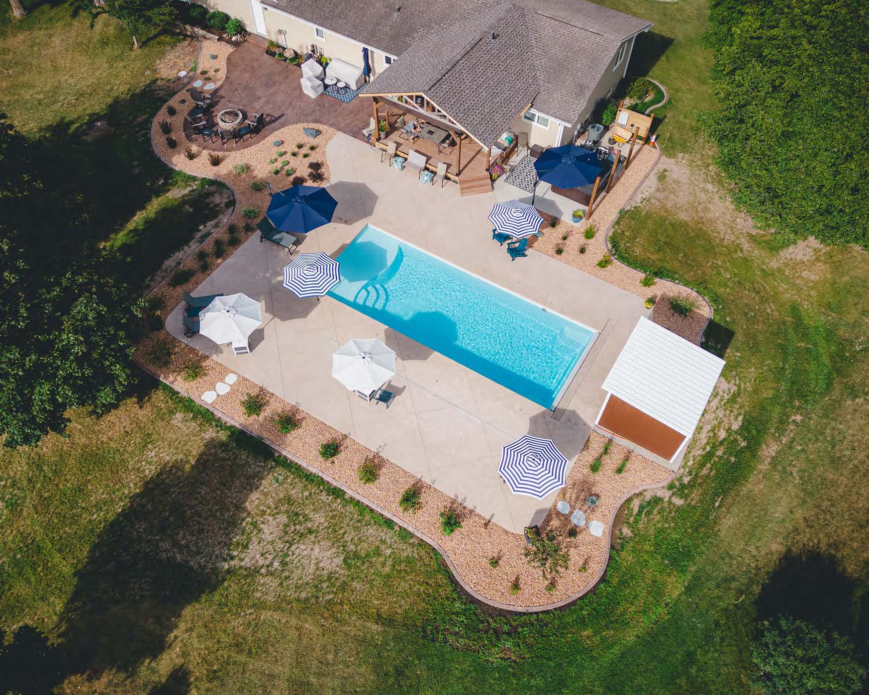 Rectangular fiberglass pool installation with patio umbrellas, fire pit, and poolside structure in a backyard near Rochester, MN, built by River Pools of Red Wing.