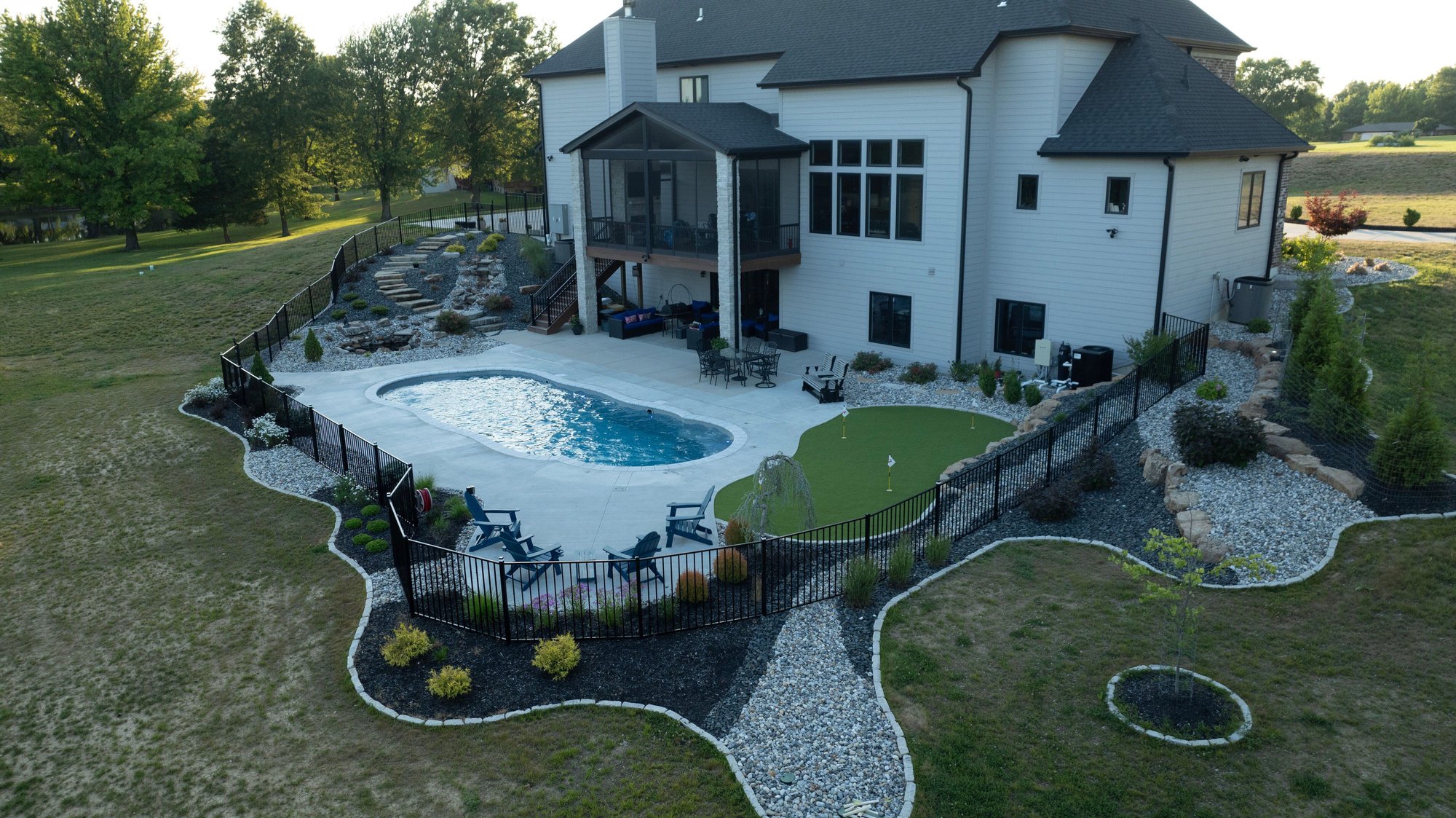 Freeform fiberglass pool installation in Arnold, MO with curved fencing, tiered landscaping, and a backyard putting green alongside the pool deck.
