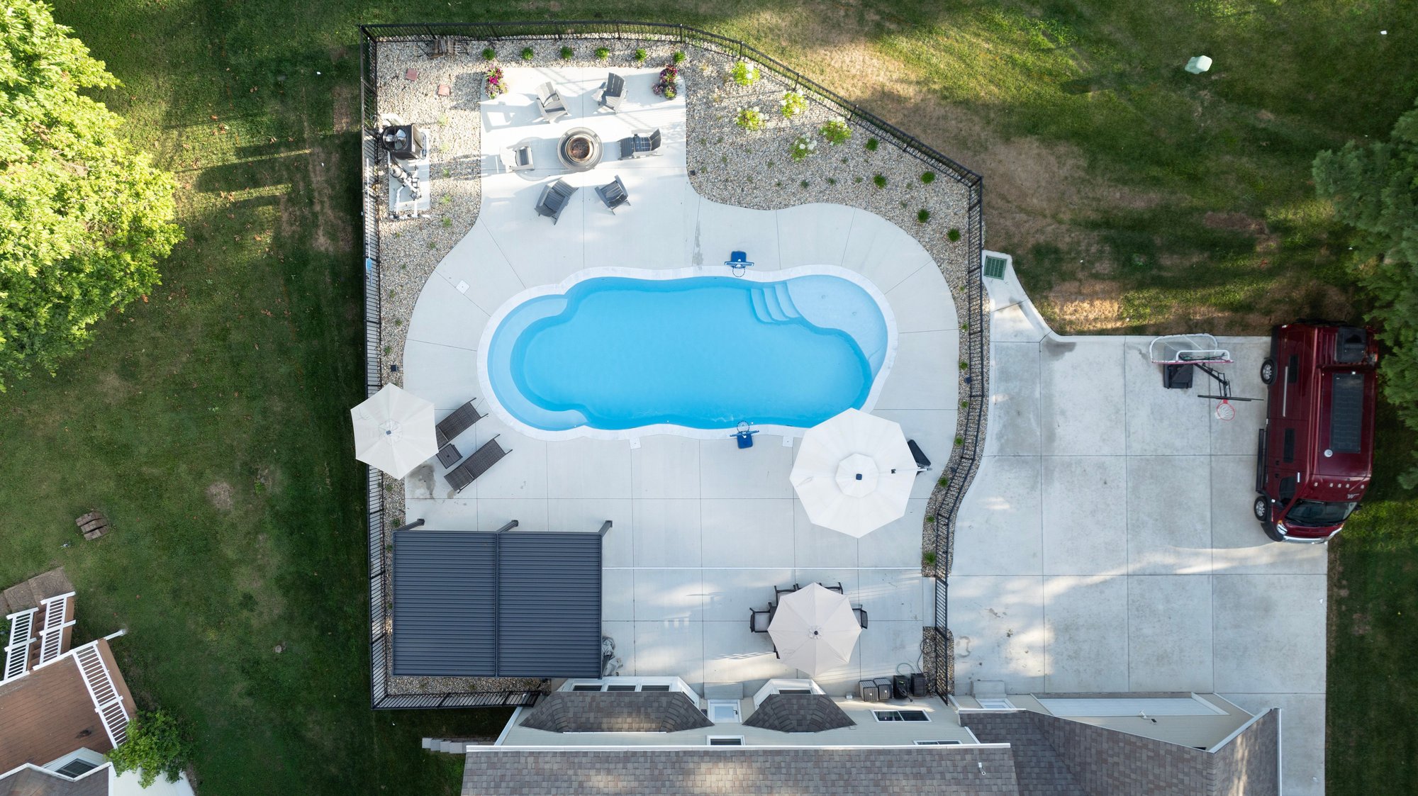 Aerial view of a fiberglass pool installation in St. Charles, MO with a freeform design, concrete pool deck, black metal fencing, shaded seating area, and nearby fire pit.