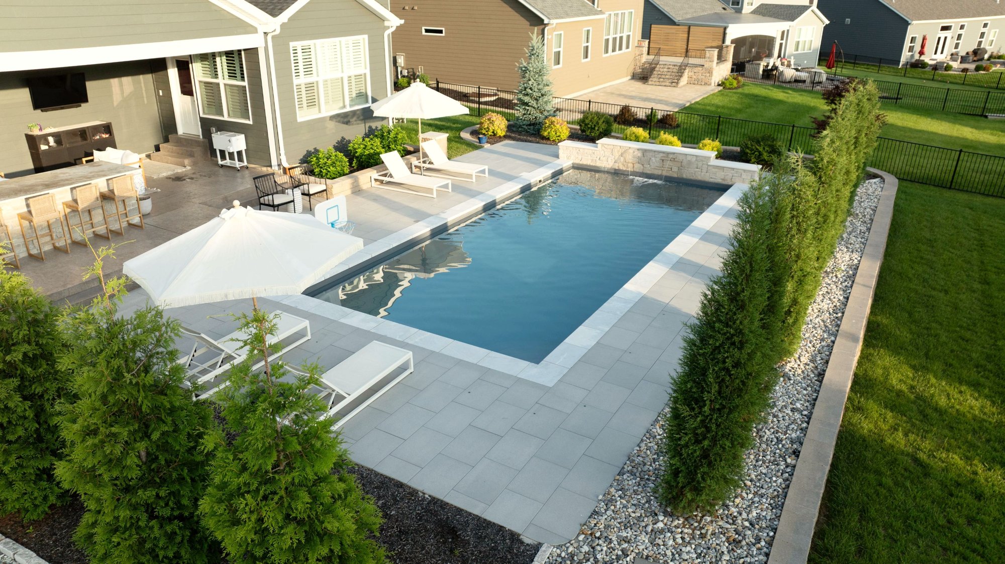 Modern fiberglass pool installation in Chesterfield, MO featuring a sleek rectangular design, black metal fencing, stone retaining wall, and private tree-lined backdrop.