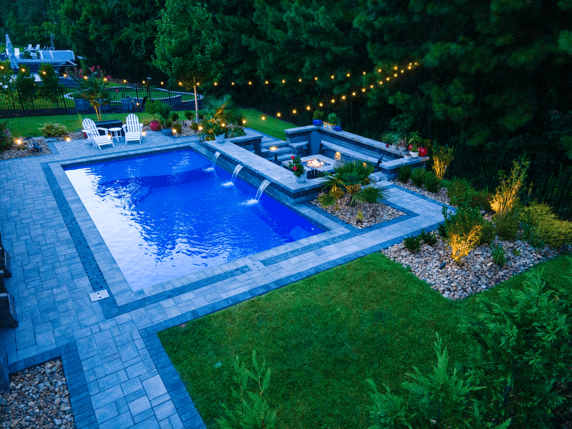 Evening view of a D Series pool with water features, glowing lights, and built-in fire pit seating area.