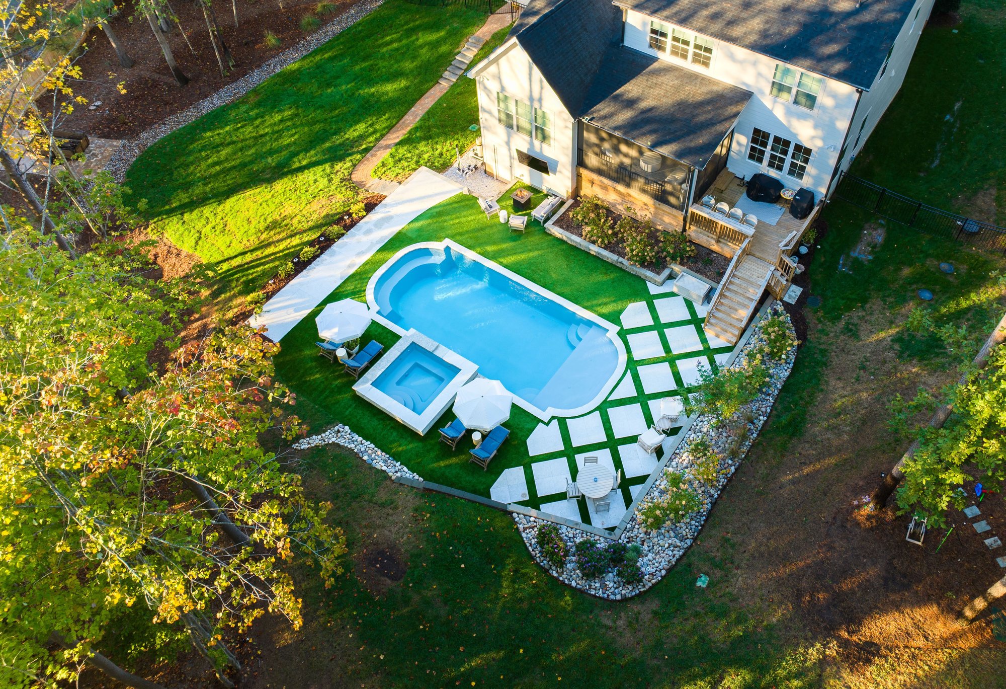 Aerial view of a modern backyard featuring a rectangular fiberglass pool with integrated hot tub, surrounded by artificial turf, white pavers, and shaded lounge areas.