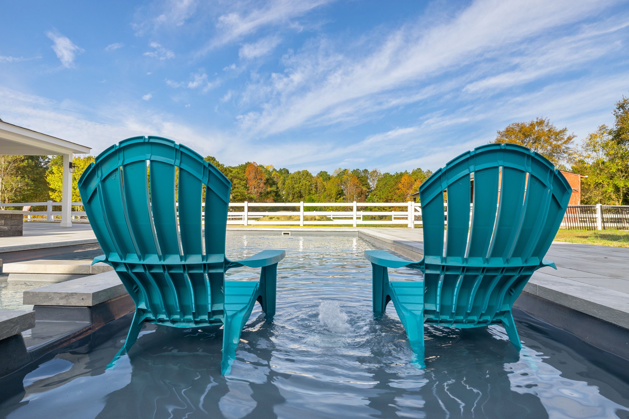 Two teal Adirondack chairs positioned on the tanning ledge of a River Pools X36, overlooking the pool with a scenic rural backdrop and white fence.