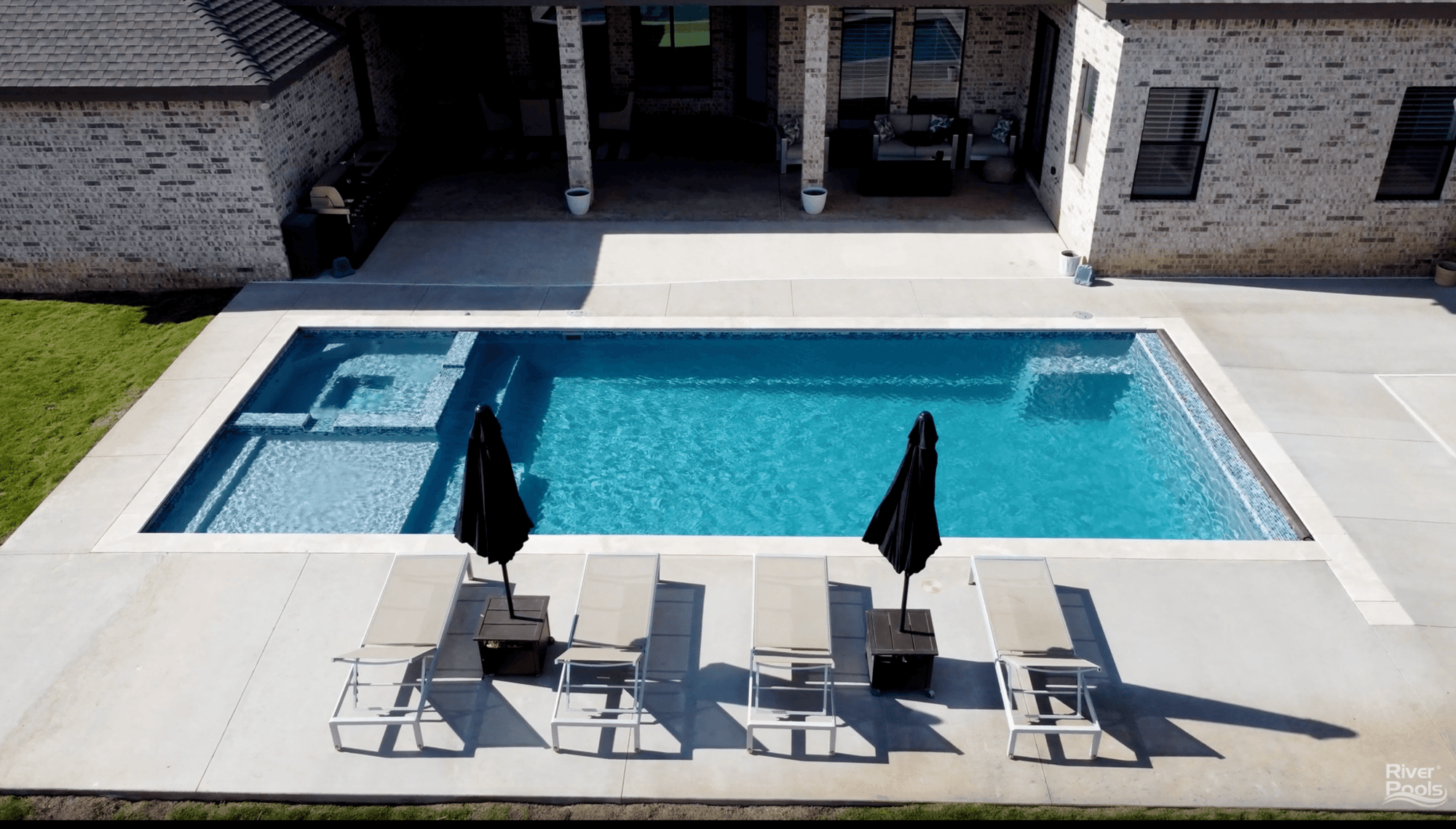 River Pools X36 fiberglass pool with built-in spa and waterline tile, surrounded by a modern concrete patio, lounge chairs, and black umbrellas beside a brick home.
