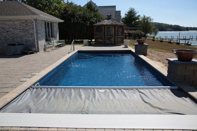 Backyard fiberglass pool with a built-in automatic safety cover partially open, surrounded by a paver patio and a wooden gazebo in the background near a waterfront.