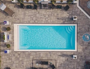 Aerial view of a River Pools Red Wing fiberglass pool installation featuring a rectangular pool with a built-in automatic cover, surrounded by a paver patio, lounge seating, and landscaping